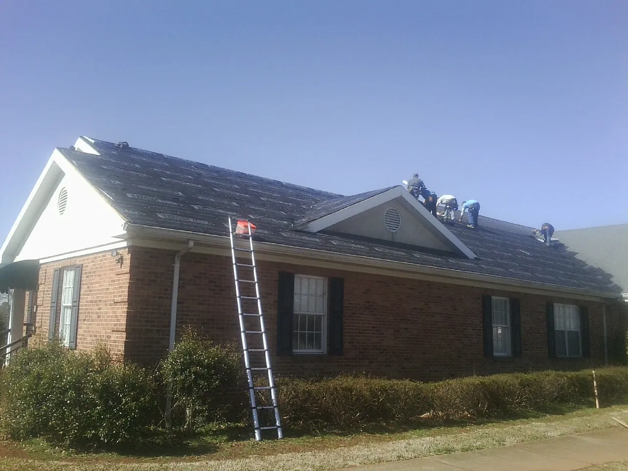 Crew tearing shingles off a brick ranch roof with a ladder set — Metal Roof Repair work by our crew in High Shoals