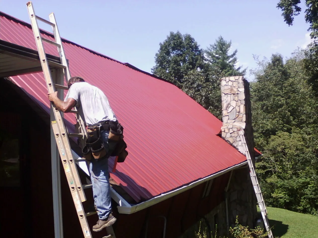 Roofer on a ladder beside a red metal roof and stone chimney — Soffit & Fascia Repair by Roofing Brothers in High Shoals